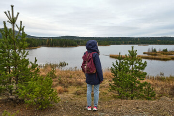 woman with backpack is standing in front of a river, forest and little island. little spruce grow near her. She put on a hoodie jacket. Solo female tourism. Lifestyle moment.