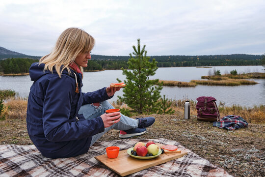woman is sitting on a plaid takes a cup of tea and a sandwich in front of a river, forest and little island. backpack, plaid, thermos near. Apples, pears, grape and sandwich lies on a green plate.