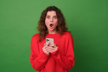 Beautiful young girl in red clothes on a green background. The girl with the phone in her hands is surprised by the discounts. The girl looks at the camera