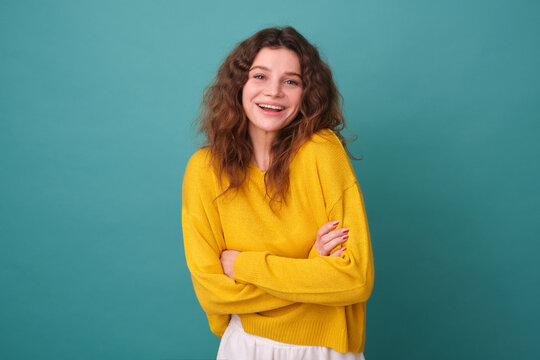 Happy joyful woman laughs happily keeps her hands folded on her chest and expresses positive emotions smiles with happiness dressed in a casual yellow jumper isolated on a blue background having fun
