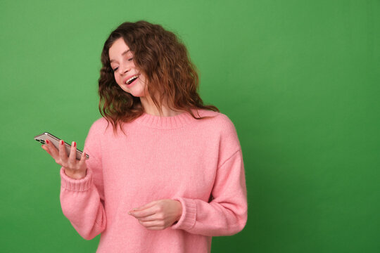 A young happy girl is smiling in a pink sweater and holding a phone on a green background
