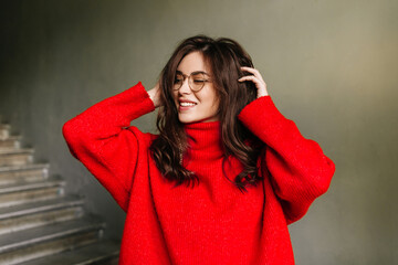 Positive brunette in high spirits touches her curly long hair. Girl in stylish oversized sweater posing on gray background