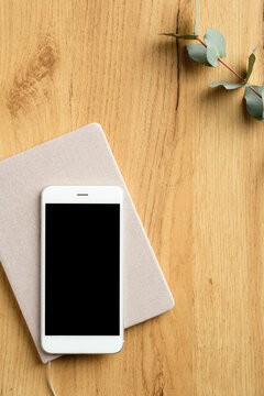 Mobile phone on wood table with paper notebook and eucalyptus leaf. Flat lay, top view home office desk.