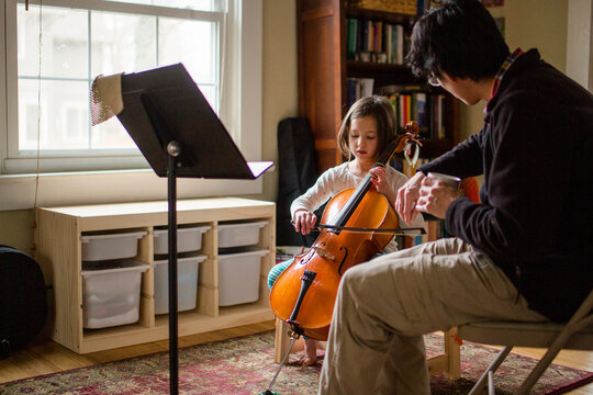 A father helps little girl practice cello at home by window