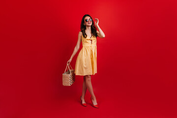 Lady in stylish summer outfit posing on red background. Curly brunette in sunglasses holds beach bag