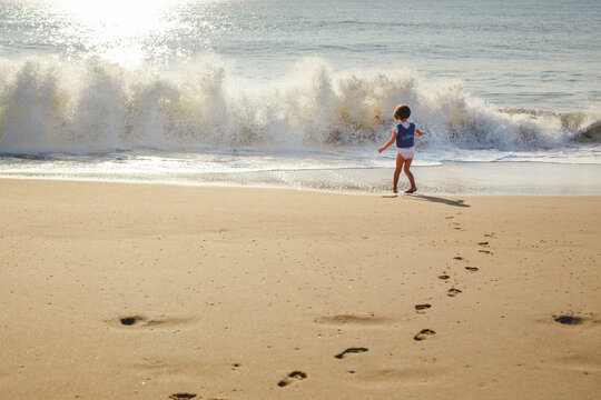 A little girl in life vest stands in front of oncoming wave at beach