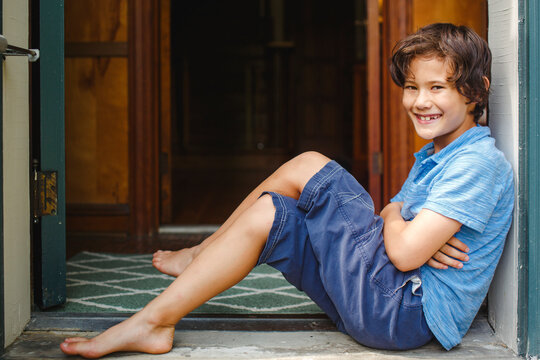 A boy with missing teeth sits smiling in a font entryway barefoot