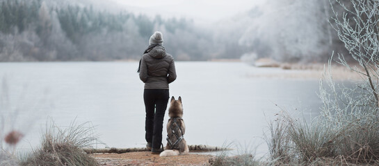 siberian husky dog sitting on the shore of a frozen lake with young female owner standing next to her in the mountains in winter panoramic