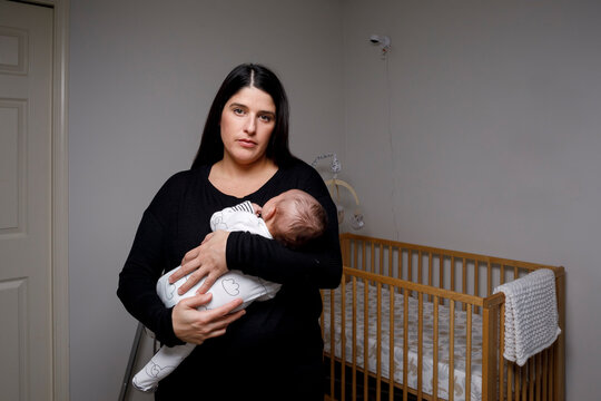 A mother holds her newborn son in arms in nursery and looks at camera