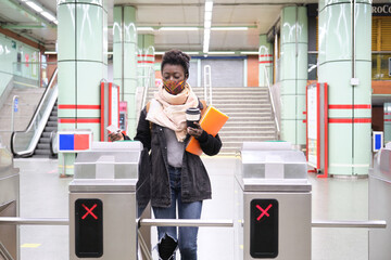 University female african student wearing protective face mask passing through the turnstiles with her transport card at the underground station. New normal in public transport.