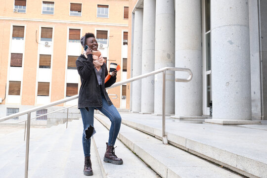 University female african student laughing and speaking on her smartphone, holding a coffee cup on the stairs of the school building on campus. College life concept.