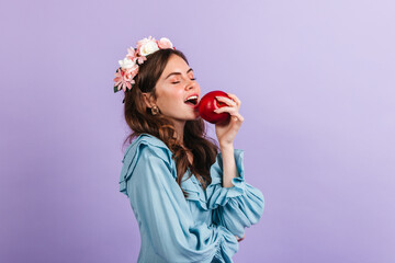 Obraz na płótnie Canvas Charming lady in a crown of flowers bites juicy apple. Portrait of girl in blue blouse on lilac background