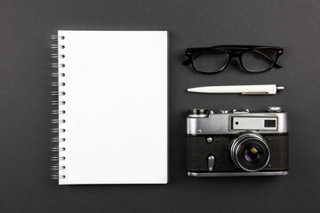 Office desk table with blank paper notebook mockup, pen, camera and glasses. Dark workspace table top with copy space. Top view flat lay.