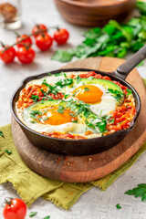 Shakshouka with avocado in a frying pan on a gray table close-up. Shakshouka of tomato sauce, eggs, spices, onions, garlic, and avocado. Tasty Maghreb style breakfast.