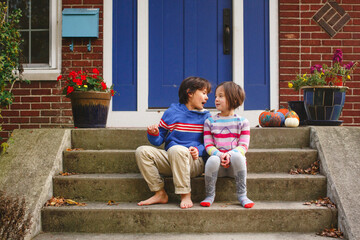 A brother and sister sit smiling together on front stoop of home