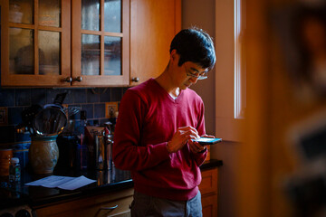 A man stands in a kitchen by window light texting on a cell phone