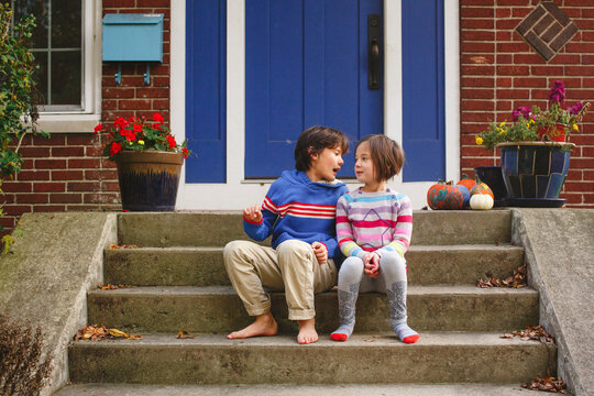 A brother and sister sit smiling together on front stoop of home