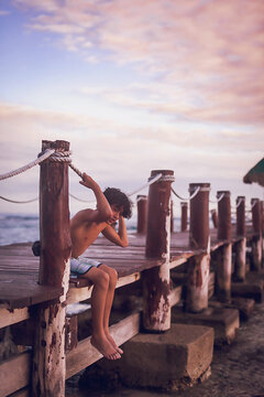 Pre teen boy sitting on a wooden pier at a tropical beach.