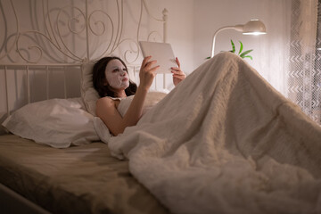 Woman with sheet mask resting on bed with tablet