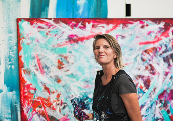 Mature artist woman looking in camera with hand full of paint colors - Masterpiece abstract oil painting in background
