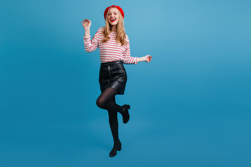 Fototapeta premium Beautiful girl in short skirt dancing on blue background. Excited well-dressed woman expressing happiness.