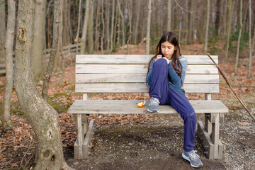 A tween girl sits alone on a bench in woods eating an orange in autumn