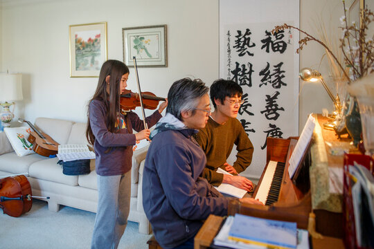 A father and uncle sit at piano while girl plays violin behind them