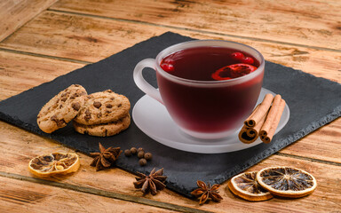 A cup of hot fruit tea or mulled wine on a slate board. Next to it is a cup of biscuits, cinnamon and dry lemon rings.