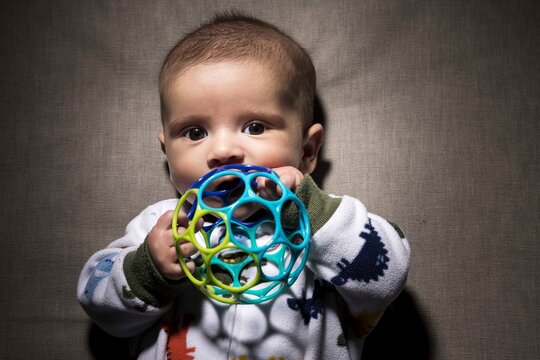 Top view of adorable newborn baby in pajama sucking colorful ball and looking at camera while lying on sofa