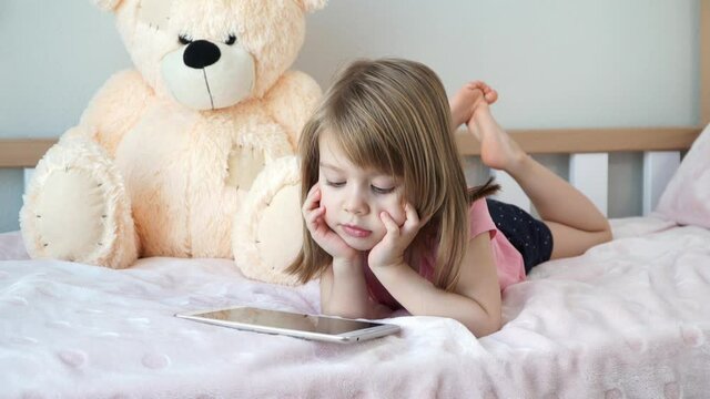 Portrait of little child girl lies at home in bed and watches digital tablet. Learning concept and education.