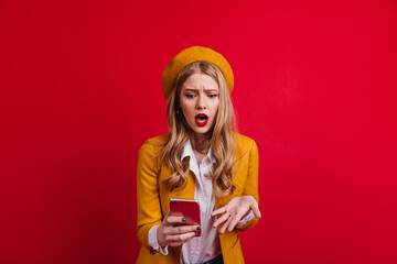 Shocked blonde woman in beret looking at smartphone. Upset french girl standing on red background with open mouth.