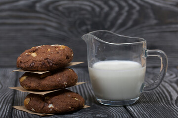 Oatmeal cookies with peanuts and chocolate. Folded in a column and laid out with paper. Nearby milk. On pine boards.