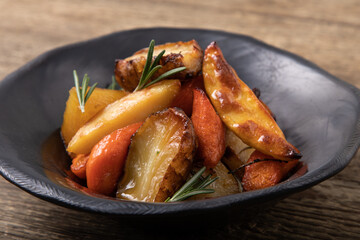 Oven baked root vegetables in handmade, black clay bowl on wooden table