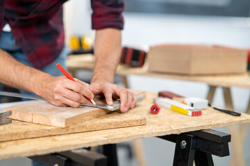 Close-up of a hardworking professional carpenter holding a angular ruler and pencil while measuring a board in a carpentry workshop. A bearded DIY enthusiast measures wood. There are a locksmith table