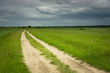 Fototapeta na wymiar Sandy road and green fields, dark clouds on the sky
