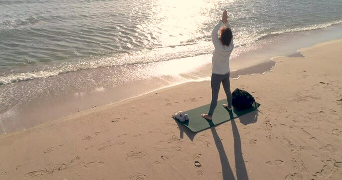 Mujer mediana edad haciendo yoga delante del mar y el sol y dron orbitando