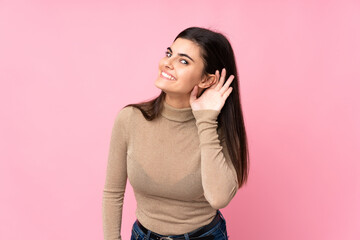 Young woman over isolated pink background listening to something by putting hand on the ear