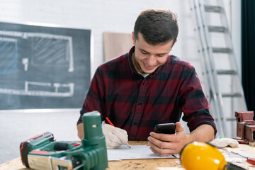 Man using cell phone and reading notes on work table. Mature carpenter working and answering phone call in his carpentry workshop.
