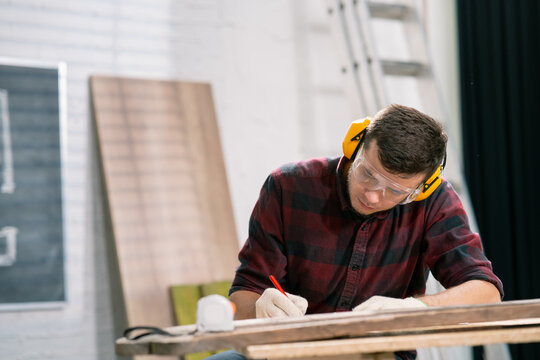 Portrait of a hardworking professional carpenter holding a pencil in a carpentry workshop. A bearded DIY enthusiast doing notes. There are a locksmith table and tools in the background.