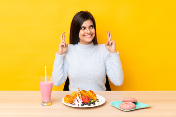 Young woman eating waffles and milkshake in a table over isolated yellow background with fingers crossing