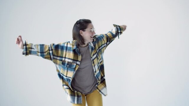 Young woman wearing round glasses, yellow pants and checkered shirt dancing alone barefoot in empty white studio