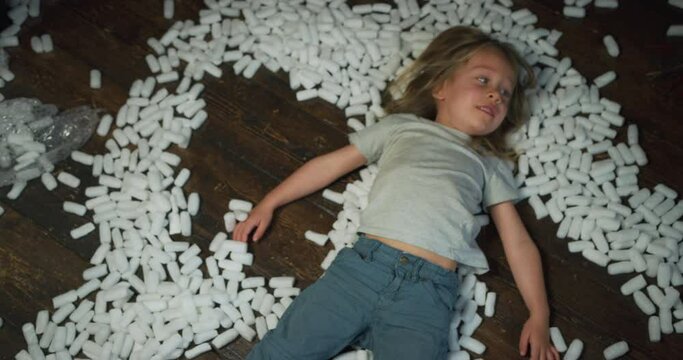 A little preschooler is making snow angels with styrofoam balls at home on the floor
