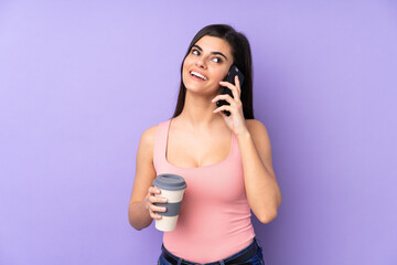 Young woman over isolated purple background holding coffee to take away and a mobile