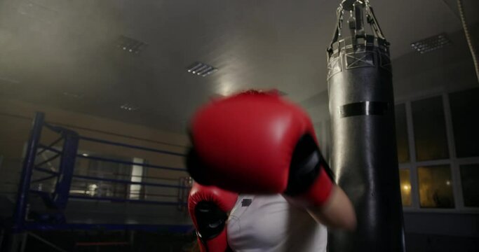 Young professional boxing girl aggressively slips into the camera lens in boxing gloves trains at night in the gym in sportswear