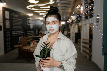 girl with a bun on her head and a clay mask on her face is holding a flower pot in a public place, in a cafe, the concept of feel at home