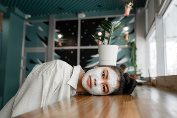 a girl with a clay mask lies on the table, and on her a pot with a growing flower, the concept of an environmentally friendly cosmetic product