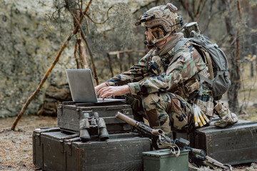 Bearded soldier in uniform sit on military transport crates, analyze data on a laptop and work out tactics at a temporary forest base. In the background, you can see a soldier protecting the base.