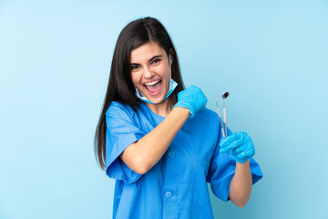 Young woman dentist holding tools over isolated blue background celebrating a victory