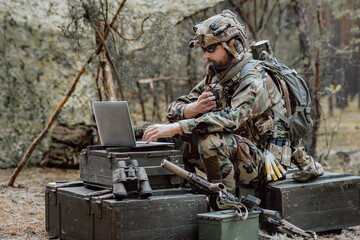 Fototapeta na wymiar Bearded soldier in uniform sit on military transport crates, analyze data on a laptop and work out tactics at a temporary forest base. In the background, you can see a soldier protecting the base.