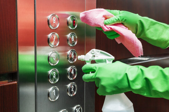 A woman uses a disinfectant spray and a wet wipe to clean the elevator button control panel.